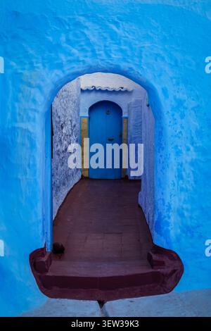 Une arche bleue éclatante encadre une ruelle étroite menant à une porte bleue fermée aux accents dorés dans la médina historique de Chefchaouen, au Maroc. Le Wal Banque D'Images
