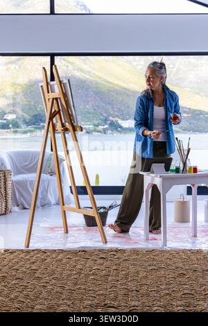 Femme asiatique senior portant du denim debout pieds nus au chevalet dans la peinture de studio avec pinceau et verre. Peintre, toile, palette, front de mer, montagnes, maison, naturel Banque D'Images