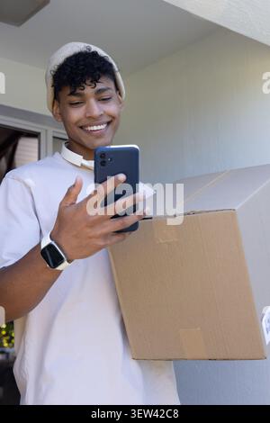 Homme afro-américain avec bonnet et smartwatch tenant le colis tout en lisant le téléphone sur le porche Banque D'Images