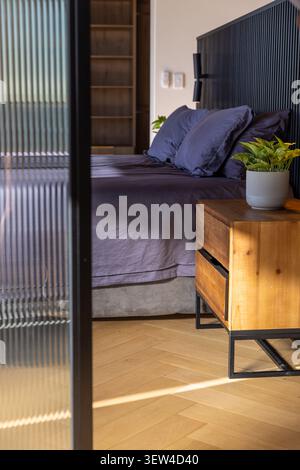 Lit reposant avec literie gris foncé à côté de la table de nuit dans la chambre, verre nervuré coulant le faisceau de soleil Banque D'Images