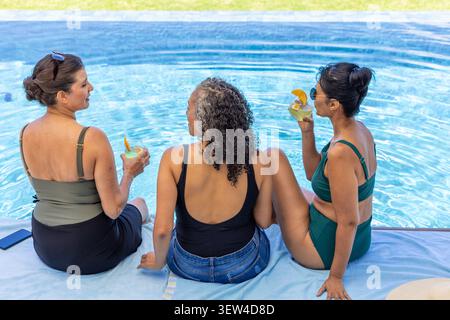 Diverses amies féminines assis sur une serviette au bord de la piscine, tenant des cocktails, tablette à proximité Banque D'Images