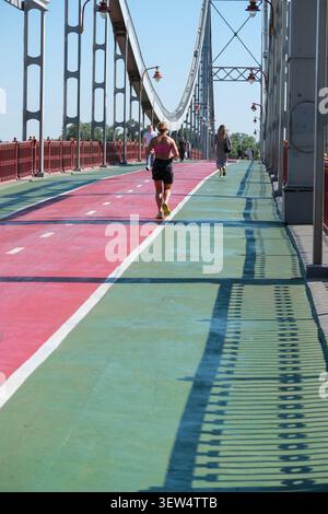 Deux personnes font du jogging le long d'un sentier de pont coloré avec la lumière du soleil tout autour. Kiev, Ukraine, 25 juin 2026 Banque D'Images