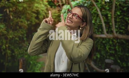 Femme avec des lunettes rouges pointe le doigt vers le haut dans la forêt, souriant et portant une chemise blanche et un blazer olive ; optimisme. Banque D'Images