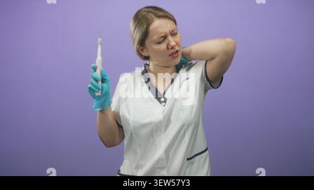 Femme dentiste portant un uniforme blanc et des gants bleus tenant une brosse à dents électrique, main sur le cou griffant dans le studio ; gêne. Banque D'Images