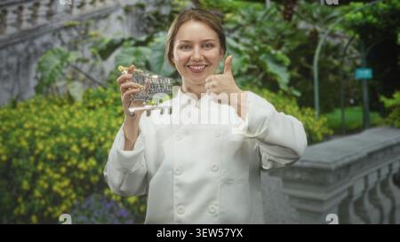 Femme chef en uniforme blanc tenant un chariot miniature et montrant les pouces vers le haut tout en se tenant près de la balustrade en pierre dans la cour du bâtiment ; optimisme Banque D'Images