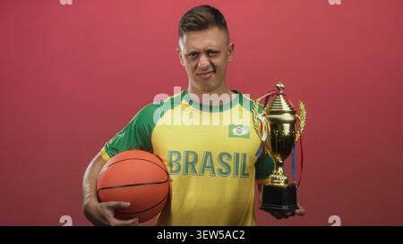 Man in brasil maillot tient basket-ball et trophée d'or avec la main droite tout en grimacant sur fond de studio rouge ; déception. Banque D'Images