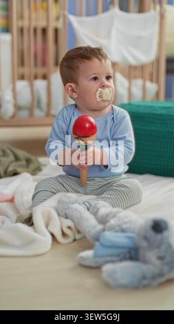 Garçon tout-petit tient maraca rouge, sucette dans la bouche, s'assoit sur une couverture près de la crèche dans le bâtiment de la pépinière ; curiosité ludique. Banque D'Images