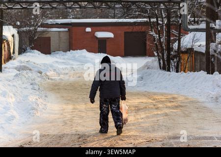Un homme seul en tenue d'hiver marche le long d'une ruelle couverte de neige transportant un sac en plastique dans la lumière du soir : Russie - 17 janvier 2026 Banque D'Images