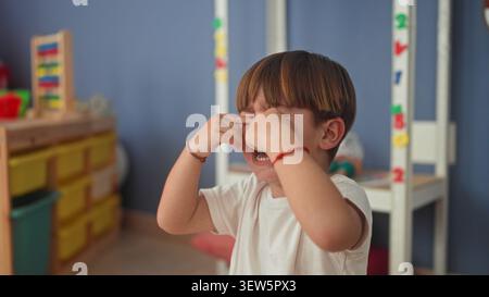 Garçon frottant les mains sur ses yeux et pleurant avec la bouche ouverte, bracelets visibles et chemise blanche dans une salle de jeux de bâtiment ; tristesse confort. Banque D'Images
