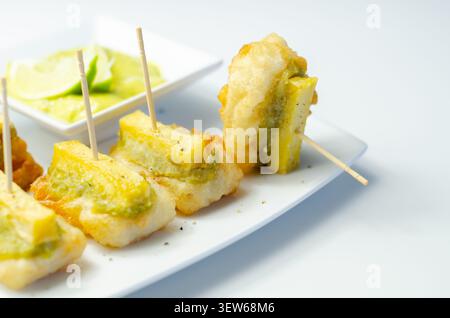Bouchées de poisson frit brun doré avec garniture aux herbes vertes sur assiette blanche. Chaque pièce percée par une brochette en bois. Garniture de citron vert frais dans un petit plat à côté. Cl Banque D'Images