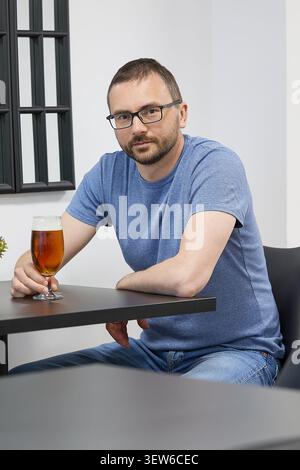 Un jeune homme avec une barbe et des verres buvant de la bière à une table dans un café Banque D'Images