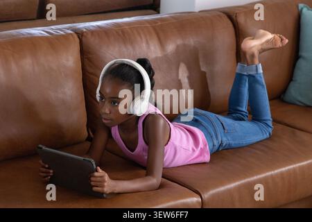 Jeunesse afro-américaine couchée sur un canapé en cuir marron à la maison portant un casque blanc et une tablette de maintien Banque D'Images