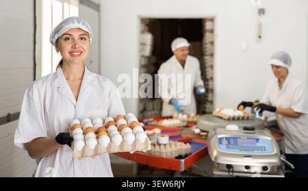 Jeune agriculteur positif en robe blanche tenant un plateau spécial avec des œufs de poulet frais après le tri et l'étiquetage à la ferme Banque D'Images