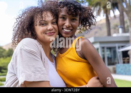 Deux amies enlacées et souriantes sur une pelouse herbeuse près d'un patio vitré avec palmiers Banque D'Images