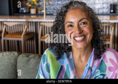 Femme afro-américaine souriante, assise sur un canapé vert dans le salon portant châle coloré, tabourets de bar Banque D'Images