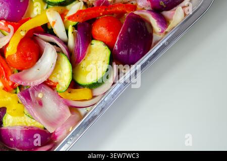 Les légumes vibrants dans un plateau métallique brillent sous une lumière vive. Les oignons rouges, les courgettes, les poivrons et les tomates cerises remplissent le cadre. Frais, coloré, an Banque D'Images