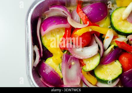 Les légumes vibrants dans un plateau métallique brillent sous une lumière vive. Les oignons rouges, les courgettes, les poivrons et les tomates cerises remplissent le cadre. Frais, coloré, an Banque D'Images