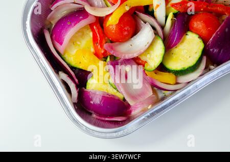 Les légumes vibrants dans un plateau métallique brillent sous une lumière vive. Les oignons rouges, les courgettes, les poivrons et les tomates cerises remplissent le cadre. Frais, coloré, an Banque D'Images