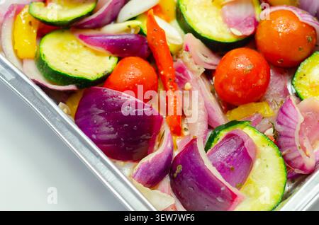 Les légumes vibrants dans un plateau métallique brillent sous une lumière vive. Les oignons rouges, les courgettes, les poivrons et les tomates cerises remplissent le cadre. Frais, coloré, an Banque D'Images