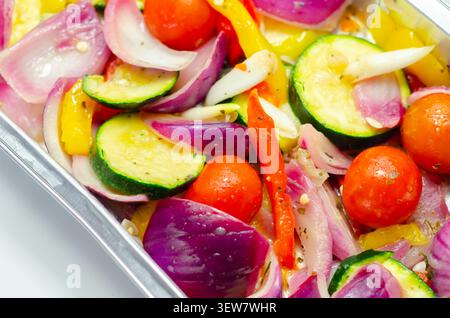Les légumes vibrants dans un plateau métallique brillent sous une lumière vive. Les oignons rouges, les courgettes, les poivrons et les tomates cerises remplissent le cadre. Frais, coloré, an Banque D'Images