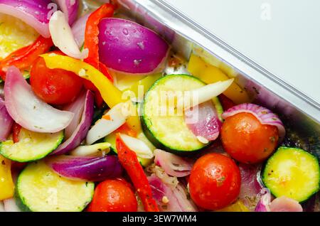 Les légumes vibrants dans un plateau métallique brillent sous une lumière vive. Les oignons rouges, les courgettes, les poivrons et les tomates cerises remplissent le cadre. Frais, coloré, an Banque D'Images