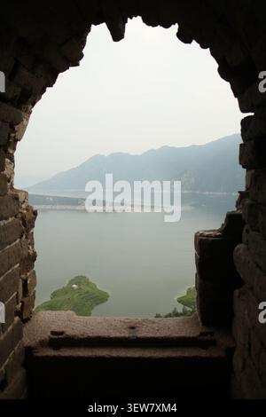 Encadré par l'arc brisé d'une tour de guet de la Grande Muraille, un paysage serein se déploie : un lac calme s'étend sous un ciel brumeux, sa surface bleu ardoise b Banque D'Images