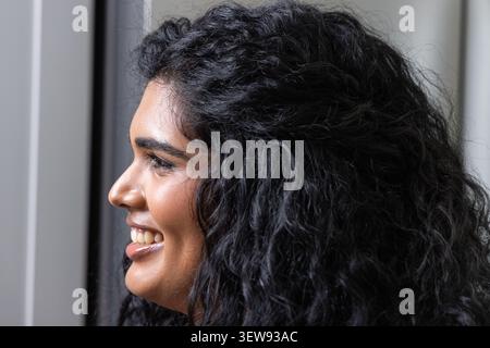 Femme indienne adulte souriante et faisant face à gauche à la fenêtre, montrant la texture des cheveux bouclés Banque D'Images