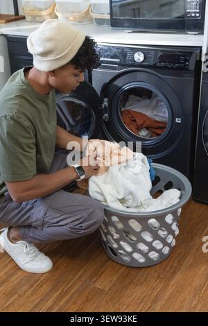 Homme afro-américain accroupi dans la laveuse de chargement de buanderie, tenant le panier gris, espace de copie Banque D'Images