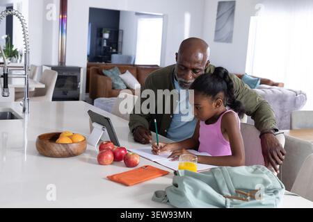 Homme afro-américain senior guidant enfant fille afro-américaine écrivant au comptoir de cuisine, crayon Banque D'Images