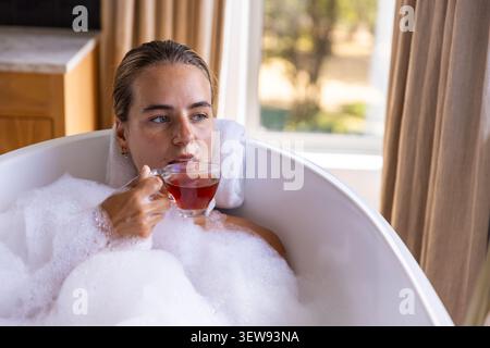 Femme dans sa vingtaine d'années s'inclinant dans une baignoire blanche autoportante avec des bulles denses tenant une tasse de verre Banque D'Images