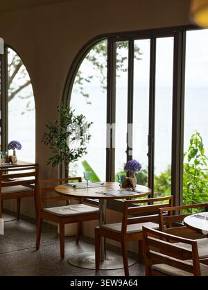 Intérieur élégant du café avec mobilier en bois, lumière naturelle douce et vue paisible sur le jardin à travers de grandes fenêtres cintrées. Banque D'Images