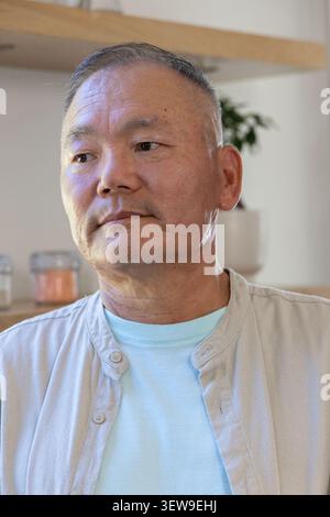 Homme asiatique senior assis dans la cuisine portant un T-shirt bleu et une surchemise beige, une usine de pots d'étagère Banque D'Images