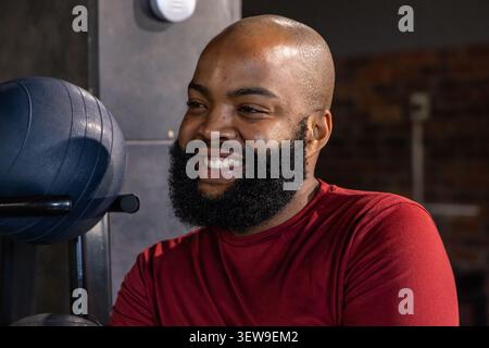 Homme afro-américain en chemise rouge à manches longues debout et souriant par rack de medicine ball au gymnase Banque D'Images