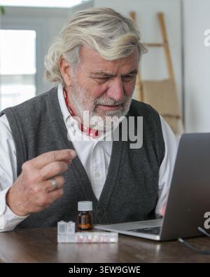 Homme senior authentique tenant des médicaments tout en utilisant un ordinateur portable lors d'une consultation en ligne à la maison. Santé, télésanté et gestion des médicaments Banque D'Images