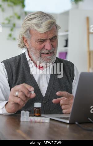 Homme senior authentique tenant des médicaments tout en utilisant un ordinateur portable lors d'une consultation en ligne à la maison. Santé, télésanté et gestion des médicaments Banque D'Images