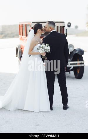 Couple romantique partage un baiser tendre à côté du véhicule vintage. Mariée porte une robe blanche fluide avec voile délicat. Le marié se tient près, habillé en b pointu Banque D'Images