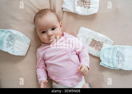 Bébé nouveau-né calme dans un body à manches longues rose couché sur une couverture douce à côté de couches propres pliées, regardant curieusement. Vue de dessus. Angle élevé Banque D'Images