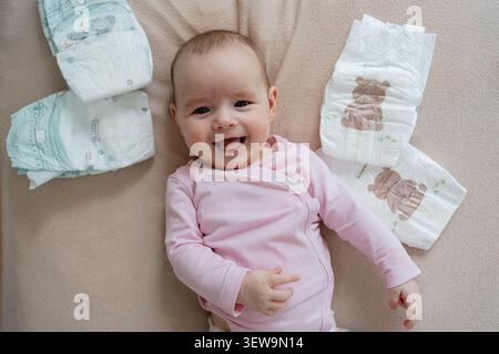 Vue aérienne d'une petite fille heureuse et souriante dans un body rose couché sur une couverture beige entourée de couches propres, regardant directement dans la caméra Banque D'Images
