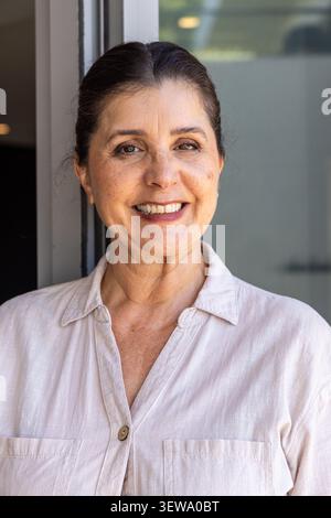 Femme senior debout dans la porte souriante, portant une chemise à col beige avec bouton, panneau de verre Banque D'Images