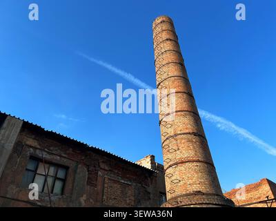 Haute cheminée d'usine de briques s'élevant au-dessus d'un bâtiment industriel abandonné contre le ciel bleu vue à angle bas Banque D'Images