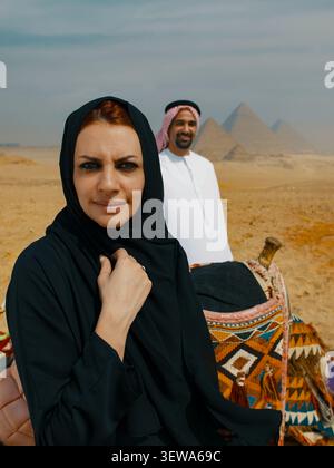 Couple arabe réfléchi sur un voyage dans le désert avec une femme sereine en hijab noir en nette mise au point et son mari souriant près d'un chameau près des pyramides de Gizeh Banque D'Images