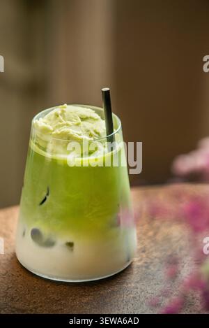 Latte matcha glacé recouvert de glace au thé vert et d'une paille noire, avec un bokeh floral rose au premier plan. Banque D'Images