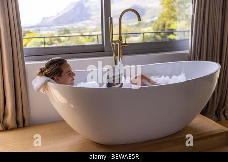 Femme couchée à la maison dans le livret de lecture de baignoire, reposant sur une serviette roulée au milieu de mousse à bulles Banque D'Images