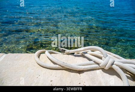 Une vue rapprochée d'une corde blanche enroulée reposant sur une surface de pierre à côté de l'eau bleue claire. La scène capture un environnement côtier tranquille. Banque D'Images