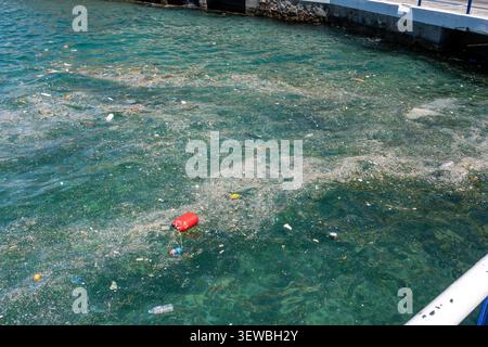Une vue surélevée de l'eau de mer claire fortement polluée par des microplastiques flottants, de l'écume de déchets, des bouteilles en plastique, et une boîte rouge près d'un harb de béton Banque D'Images