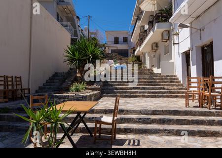 Une vue panoramique sur une place tranquille en Crète avec des chaises en bois rustiques et des tables d'un restaurant en plein air situé sur de larges escaliers de pierre sous un blu clair Banque D'Images