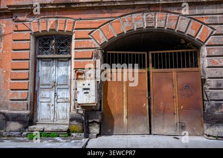Ancienne façade en briques avec porte voûtée. Portails en bois vieilli avec barres métalliques. Boîte aux lettres rouillée à côté de la porte décolorée. De petites plantes vertes poussent à la base. Urbain Banque D'Images