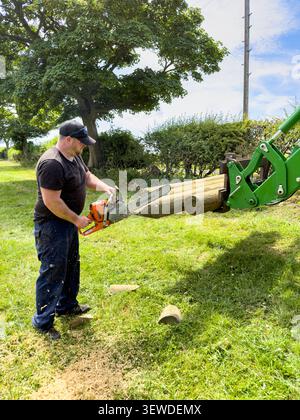 Homme coupant des poteaux de clôture en bois avec une tronçonneuse Stihl et un chargeur de tracteur John Deere vert dans un cadre rural Banque D'Images