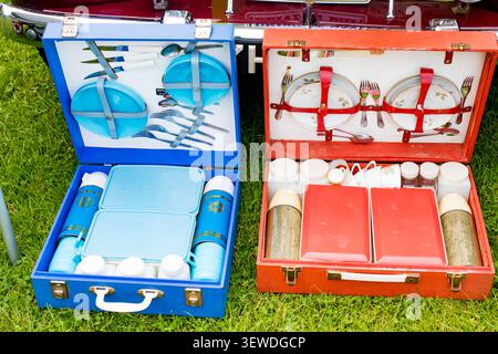 Deux paniers de pique-nique vintage ouverts avec des ensembles de salle à manger en plastique bleu et rouge sur la pelouse verte à côté d'une voiture classique Banque D'Images