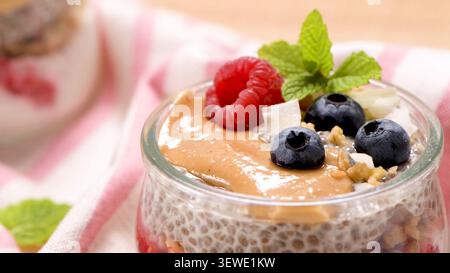 Un gros plan d'un pot rempli de pudding crémeux aux graines de chia, garni de beurre de noix lisse, de framboises fraîches, de bleuets dosés, de noix de coco râpée et d'un Banque D'Images